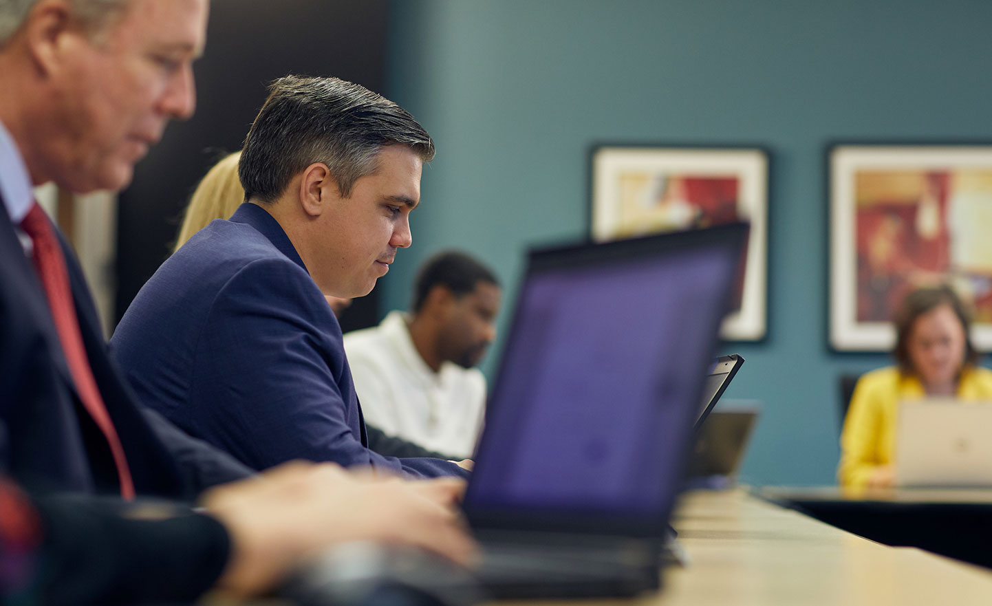 Access-Sciences-Acquisition,-Mergers,-and-Divestitures-Capabilities-and-Services People work on laptops at a conference table, centered on a man in a blue suit.