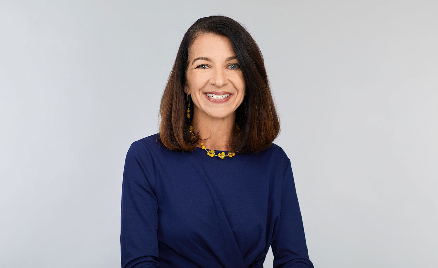 A woman in a navy top and yellow necklace smiles against a light gray background.