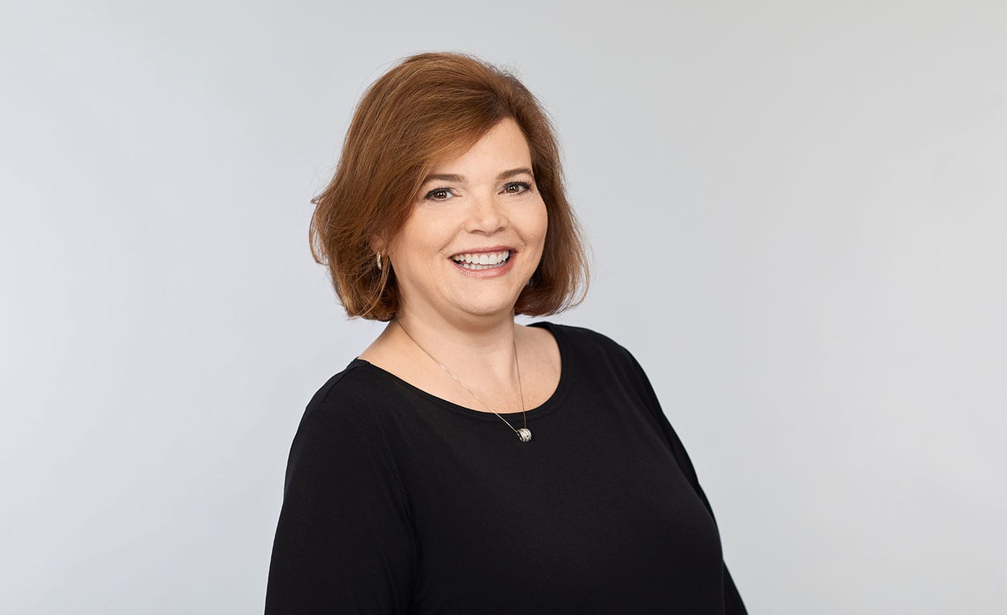 Sondar Ludwick with shoulder-length brown hair in a black top and necklace stands against a light gray background.