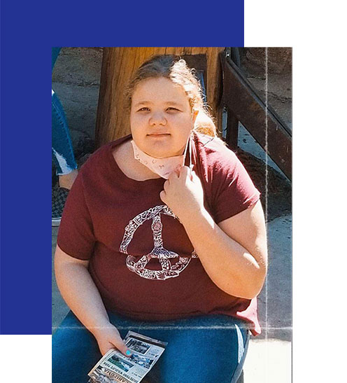 Access-Sciences-Blog-Supporting-Working-Moms-SARAH-BYBEE-2 A blonde-haired girl sits outdoors in a maroon peace-sign shirt, holding a mask and papers.