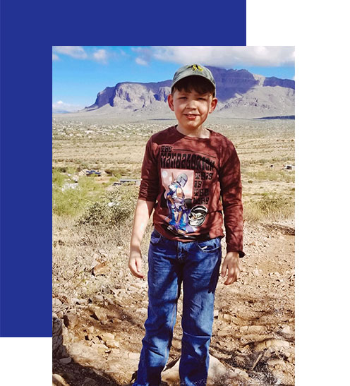 Access-Sciences-Blog-Supporting-Working-Moms-SARAH-BYBEE A young boy in a cap and maroon T-shirt stands on a rocky desert trail, with mountains and sky behind him.