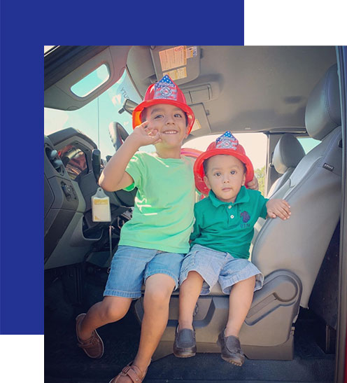 Access-Sciences-Blog-Supporting-Working-Moms-Sapana-2 Two kids in firefighter hats smile in a vehicle, capturing the joy of supporting working moms with family fun.