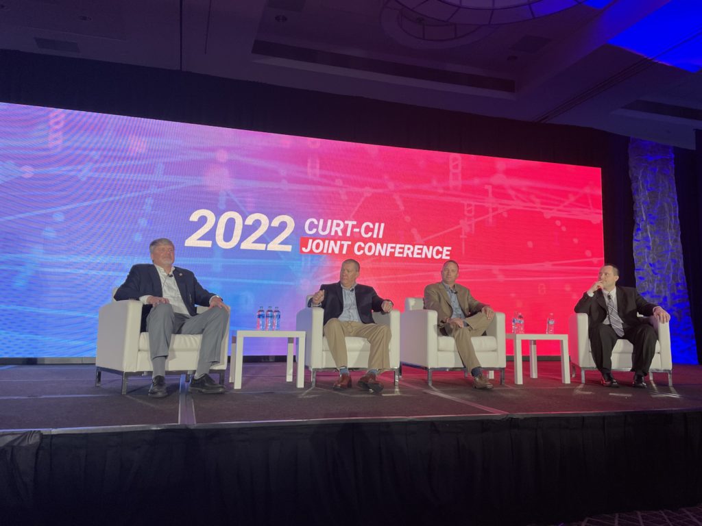 Access Sciences Blog CURT Four men in suits sit onstage in white chairs, discussing capital projects, with "2022 CURT-CII Joint Conference" displayed behind them.