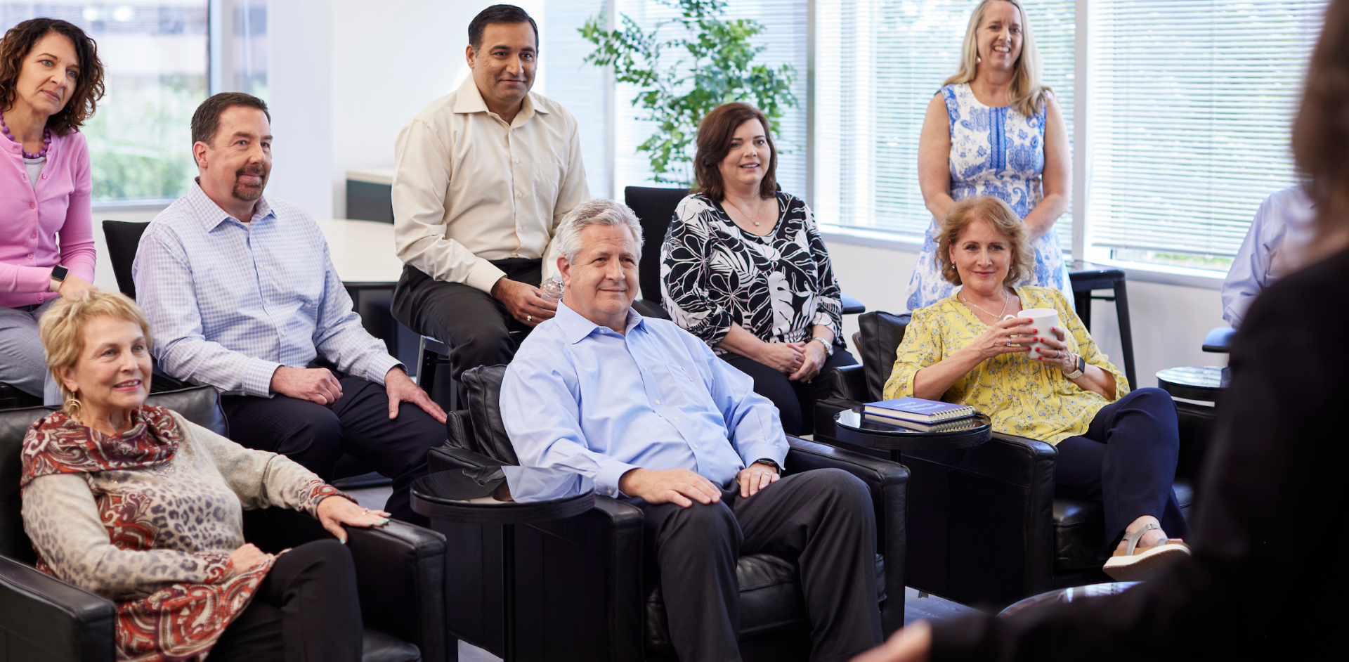 A group in a bright office listens to a speaker, with notebooks, coffee, and a welcoming setting.