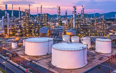 An oil refinery at sunset, featuring large white storage tanks in the foreground and numerous industrial towers and pipes in the background, with hills and a colorful sky reflecting the importance of data governance in information management.