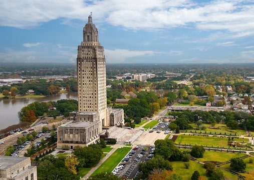 A tall, art deco-style skyscraper stands amid green trees and lawns, with roads and cars nearby under a partly cloudy sky. The building is the Louisiana State Capitol in Baton Rouge, where data governance shapes effective information management for the state.
