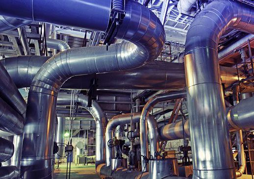 Large, shiny metal pipes and ducts run through this chemicals industry facility, reflecting light. Valves, fittings, and structural supports are visible among the complex network of pipes inside the spacious building.
