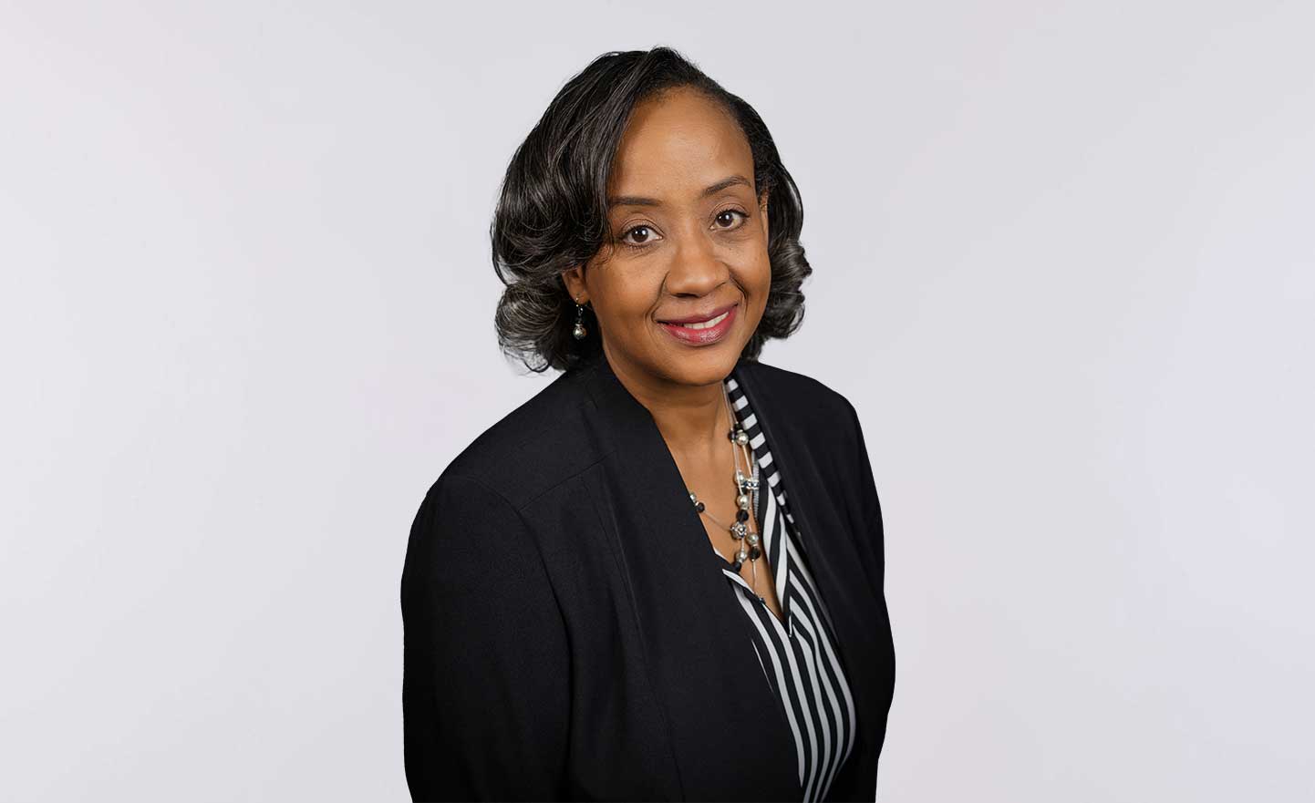 A woman with short, wavy dark hair smiles at the camera. She is wearing a black blazer over a black and white striped blouse and a statement necklace, with a plain light background.