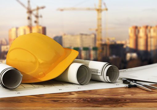 A yellow construction helmet rests on rolled-up blueprints and a wooden table, hinting at the vital role of the chemicals industry, with cranes and buildings blurred in the background.