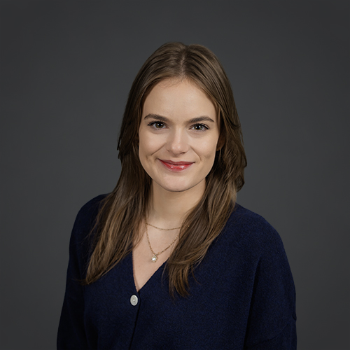 A woman with long brown hair wearing a dark blue sweater and a gold necklace, smiling at the camera against a plain dark gray background.