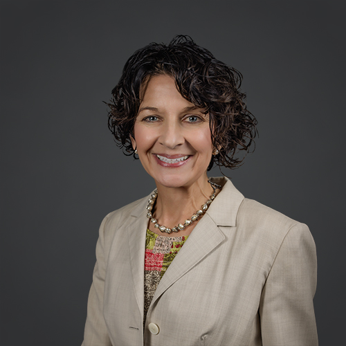 A woman with short, curly dark hair smiles at the camera. She is wearing a light beige blazer, a colorful top, and a beaded necklace. The background is plain and dark gray.