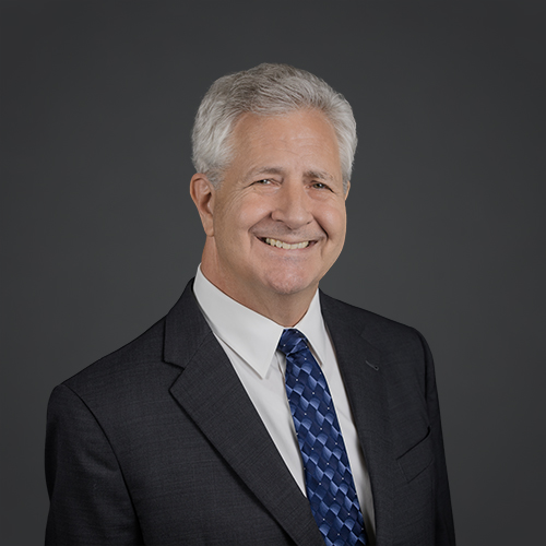 Smiling older man with short gray hair wearing a dark suit, white shirt, and blue patterned tie, posing against a plain dark gray background.