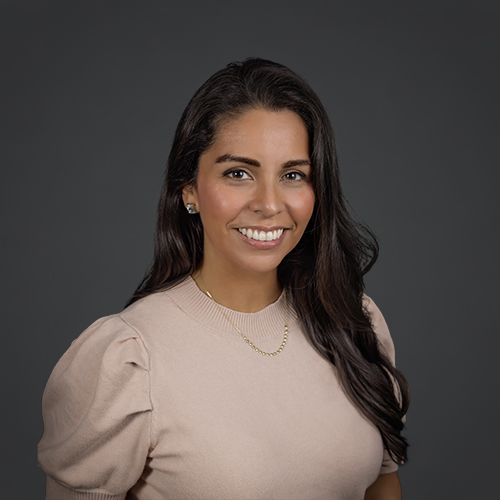 A woman with long dark hair and a beige puff-sleeve top smiles at the camera against a plain dark gray background. She wears a gold necklace and stud earrings.