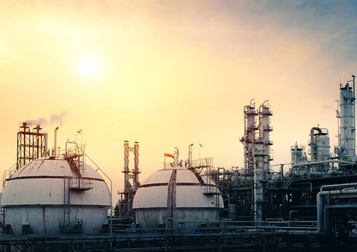 Industrial facility housing large spherical storage tanks and tall distillation towers, part of the power utilities sector, emits smoke under a hazy sky during sunset or sunrise.