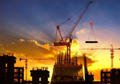 A construction site at sunset, with several cranes—hallmarks of leading E&C companies—lifting materials and silhouetted buildings in progress against a dramatic orange and yellow sky.