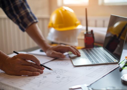 A person from E&C companies reviews architectural blueprints on a desk with a laptop, pencil, and radio; a yellow safety helmet sits in the background.
