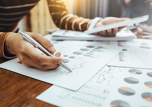 A person at an E&C company reviews and analyzes financial charts and graphs on printed documents, using a pen to point at data while holding a tablet. Sunlight streams in, illuminating the workspace.