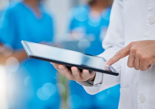 A person in a white lab coat uses a tablet device, highlighting the integration of life sciences technology, with medical staff in blue scrubs blurred in the background, suggesting a healthcare or hospital setting.