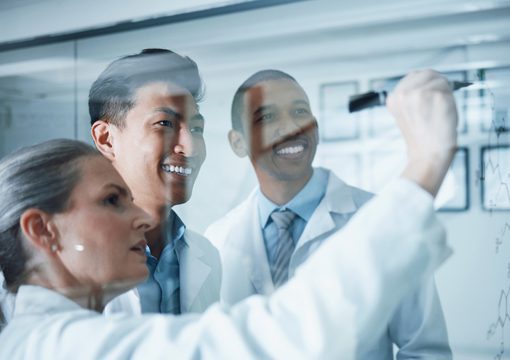 Three scientists in lab coats smiling and collaborating as one writes life sciences diagrams and graphs on a transparent board, in a bright, modern laboratory setting.
