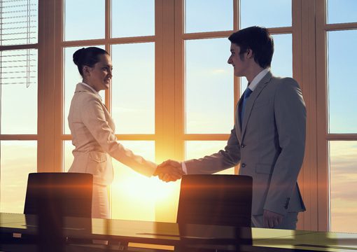 A woman and a man in business attire shake hands in an office with large windows, as the sun sets or rises in the background, creating a warm glow—signaling a promising partnership in the life sciences industry.
