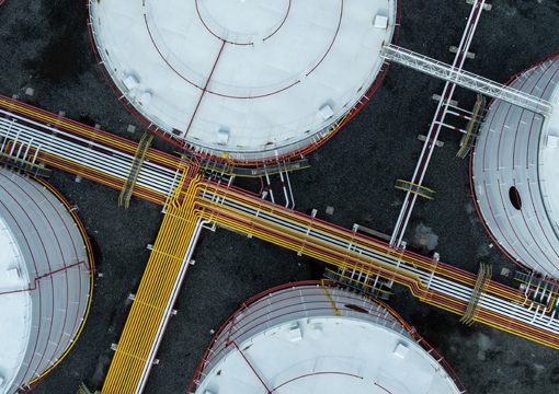 Aerial view of large round industrial storage tanks connected by yellow, red, and white pipelines, set on a dark gravel surface. The tanks and pipes form a geometric pattern typical of the chemicals industry.