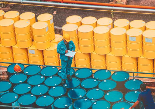 A worker in a blue uniform and yellow helmet inspects blue barrels at a chemicals industry site, taking notes on a clipboard, with stacked yellow barrels in the background.