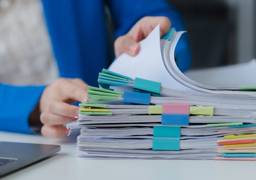 A person wearing a blue jacket sorts through a stack of documents clipped with colorful binder clips, likely reviewing important reports related to the chemicals industry, with a laptop partially visible in the foreground.