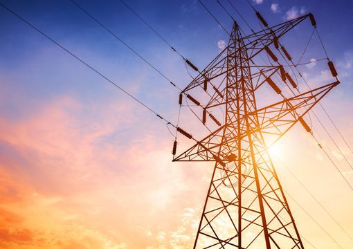 High-voltage power lines and a metal transmission tower, essential to power utilities, are silhouetted against a colorful sky at sunset, with the sun shining brightly in the background.