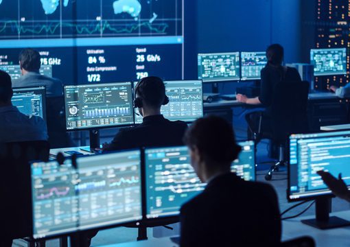 People work at computer stations in a dark control room filled with monitors displaying data, graphs, and maps for power utilities. Large screens show world maps and analytics, creating a focused, high-tech atmosphere.