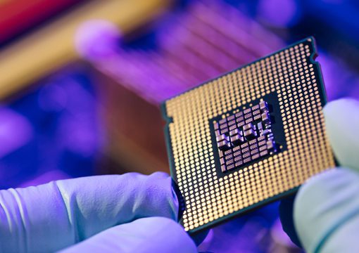A close-up of a person wearing blue gloves holding a computer processor chip, showing the gold contact points on the underside, with a blurred background of semiconductor-fabrication-plants and computer components.