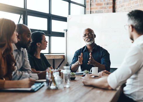 A group of five people sit around a table in a modern office, listening to an older man speaking and gesturing with his hands, with a whiteboard in the background.