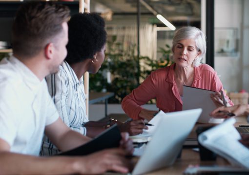 Three people sit at a table with laptops and documents, engaged in discussion. An older woman in a pink shirt speaks about information strategy, while two colleagues listen attentively in a modern office setting.