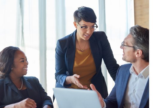 Three business professionals have a discussion in a bright office. A woman in the center speaks about managed services while gesturing, as a man and another woman nearby listen attentively. A laptop is open in front of them.