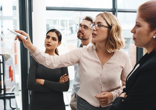 Four people in business attire stand in an office, focused on a woman who is pointing at notes on a glass wall with a marker during a team meeting on managed services or a brainstorming session.