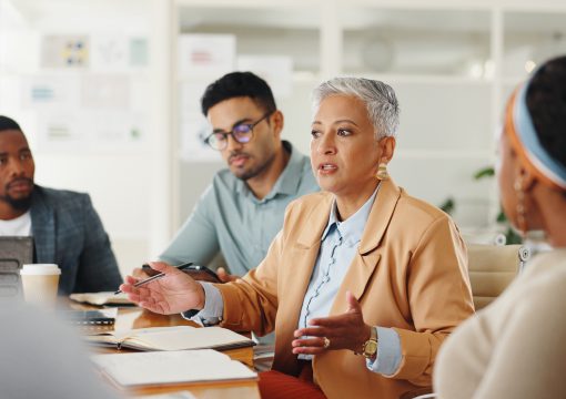 A group of professionals sit around a table in an office. A woman with short gray hair and a tan blazer discusses managed services, gesturing with her hands, while others listen attentively. Notebooks, laptops, and a coffee cup are visible on the table.