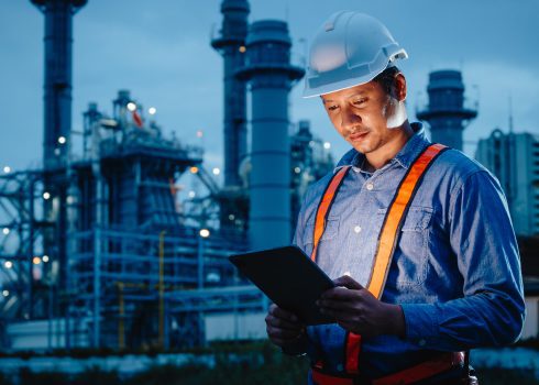 A worker wearing a white hard hat and orange safety harness uses a tablet at an industrial facility at dusk, highlighting the commitment to safety and efficiency championed by Access Sciences, an information management and governance services firm.