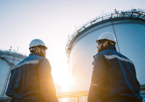 Two workers wearing hard hats and safety jackets stand outside near large industrial storage tanks at sunset, discussing technology governance or inspecting the facility as part of change management efforts.