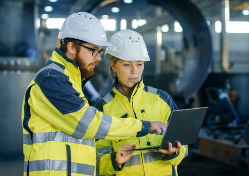 Two engineers in high-visibility jackets and white hard hats stand in an industrial setting, discussing change management strategies as they review information on a laptop that one of them is holding.