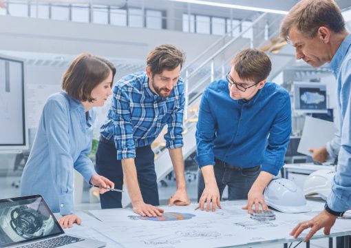 Four people in an office stand around a table, closely examining blueprints and diagrams. One woman gestures with a pen while three men discuss technology governance, collaborating on a technical project. A laptop and hard hat are visible on the table.