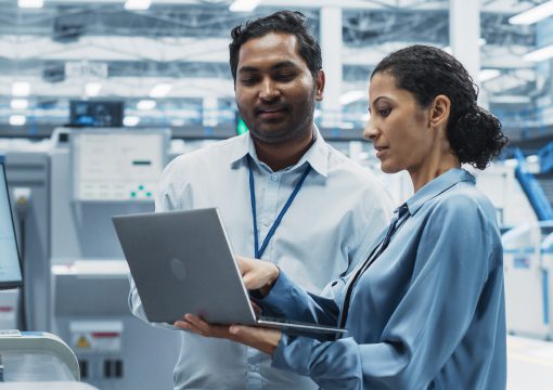 Two professionals wearing ID badges review data on a laptop together in a modern industrial facility, surrounded by machinery—demonstrating efficient operations supported by expert information governance services.