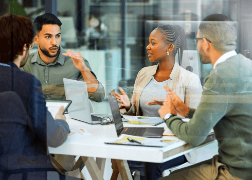 Four people are sitting around a table in an office, discussing information governance services. Laptops and papers are on the table, and everyone appears engaged in conversation. The scene is viewed through a glass wall.