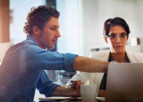 A man and a woman sit at a desk, looking at a laptop screen. The man, from Access Sciences information management and governance services firm, points at the laptop while the woman listens attentively. A coffee cup rests on the table.