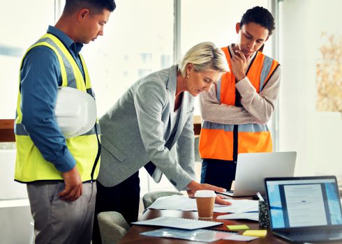 Three people, two in safety vests and one in business attire, review documents on a table with a laptop, coffee, and paperwork—highlighting the collaborative nature of information management services across industries—in an office with large windows. One holds a hard hat.