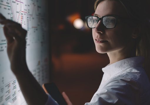 A woman wearing glasses and a white shirt points at data on a brightly lit board while holding a smartphone, concentrating on information in a dimly lit environment at Access Sciences, an information management and governance services firm.