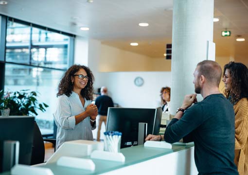 A woman stands and smiles while talking to two people at a reception desk in a modern office or clinic setting, where information governance services help ensure secure management of data. Computers, office supplies, and other staff are visible in the background.