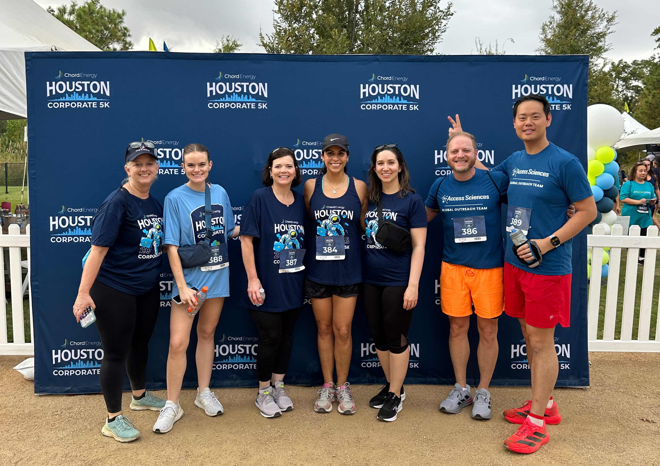 Seven people in athletic wear stand smiling together in front of a Houston Corporate 5K event backdrop, representing their team’s Global Outreach. Some wear medals and race bibs. The group is outdoors, with one person flashing a peace sign playfully.