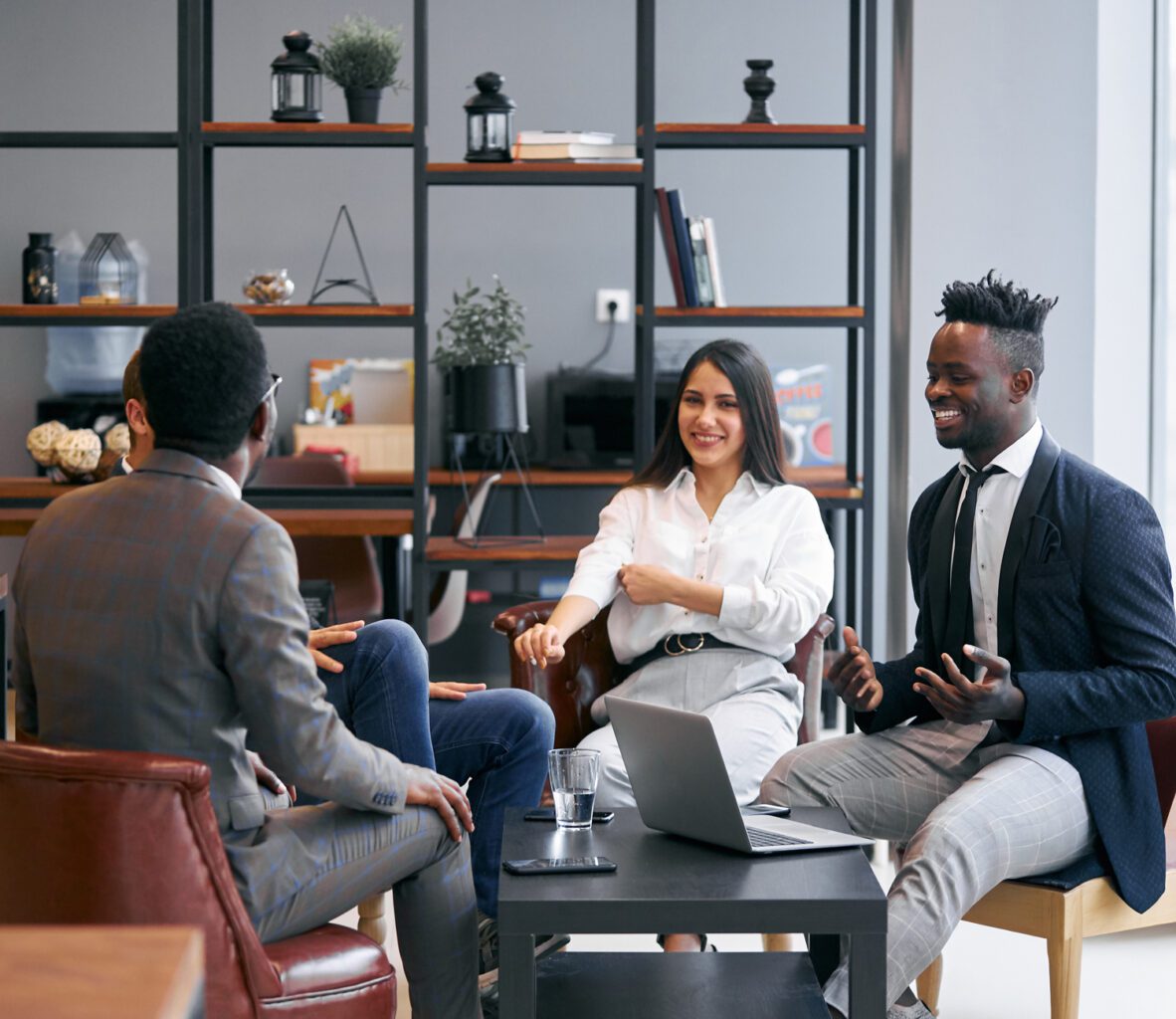 Four professionally dressed people sit together in a modern office, smiling and having an engaging discussion around a coffee table with a laptop and glass of water. Shelves with decor are in the background.