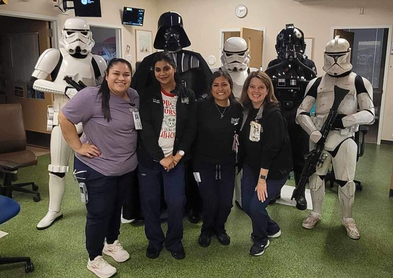 Four smiling healthcare workers pose together indoors, while people dressed as Star Wars characters—including Darth Vader, Stormtroopers, and others—stand behind them. The scene is cheerful and playful.