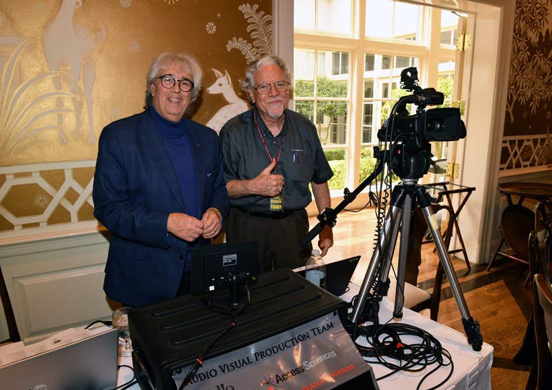 Two smiling older men stand behind audiovisual equipment and a large camera indoors. One gives a thumbs up. Sunlight streams through large windows behind them. A sign reads AUDIO VISUAL PRODUCTION TEAM.