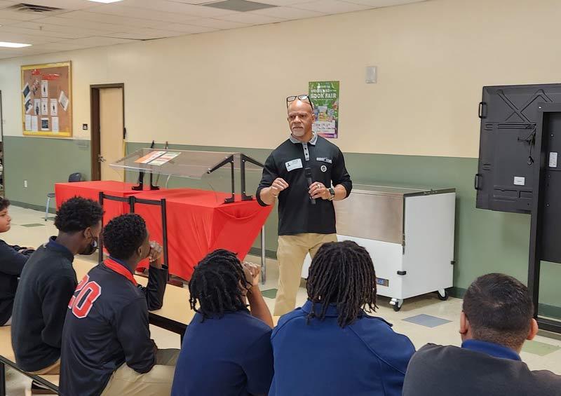 A man stands and speaks to a group of seated students in a classroom or cafeteria setting. The students listen attentively; some wear uniforms. Behind the man are tables covered with red cloths and wall posters.